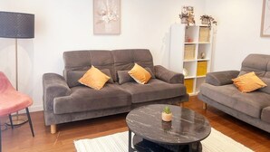 Un salon avec deux canapés gris, une table basse ronde noire, un fauteuil rose et une bibliothèque agrémentée d’objets décoratifs.