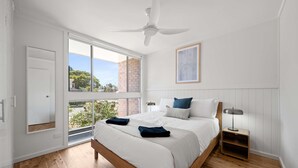 A bedroom with a large window, a ceiling fan, a bed with pillows, a bedside table, and a framed picture on the wall. - Prickly Pear Stays in Merewether - Ridge St (Newcastle)