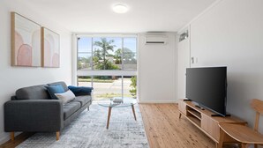 A modern living room with a grey sofa, a glass coffee table, a flat-screen TV on a wooden stand, and a large window with a view of greenery and buildings. - Prickly Pear Stays in Merewether - Ridge St (Newcastle)