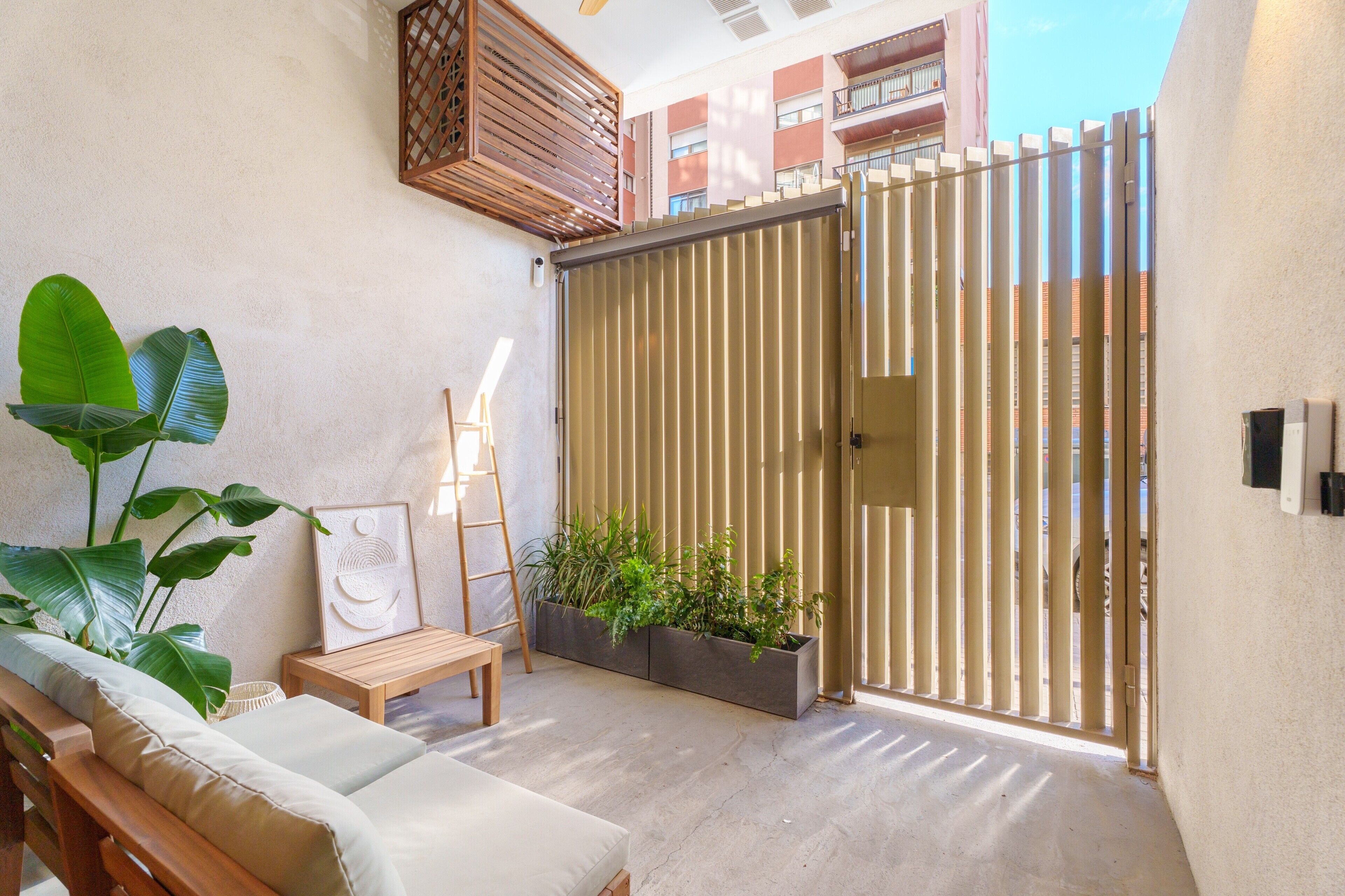 Une terrasse ensoleillée avec un banc en bois, une œuvre d’art encadrée et un jardinier contenant des plantes vertes.