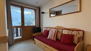 A wooden sofa with red and patterned cushions, a television on a wooden stand, and a large window with a view of snow-covered mountains. - Cosy apartment ski-in/ski-out (Les Allues)
