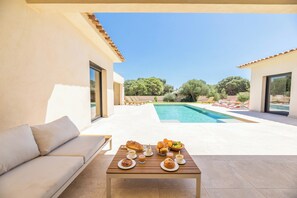 A poolside area with a table set for breakfast, a sofa, and a view of a swimming pool and trees. - Villa Anna - 10 minutes from the beach (Bonifacio)