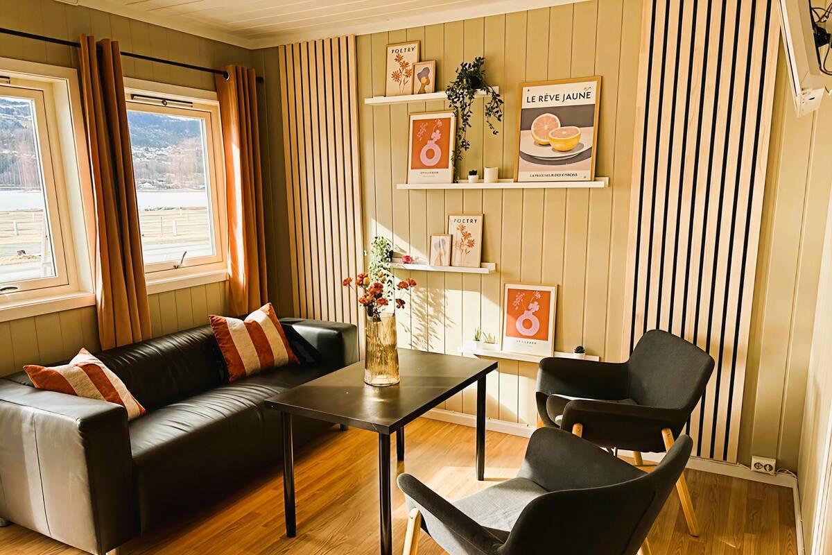 A modern living room with a sofa, a coffee table, and two chairs. There are wooden shelves with framed pictures and a window with curtains.