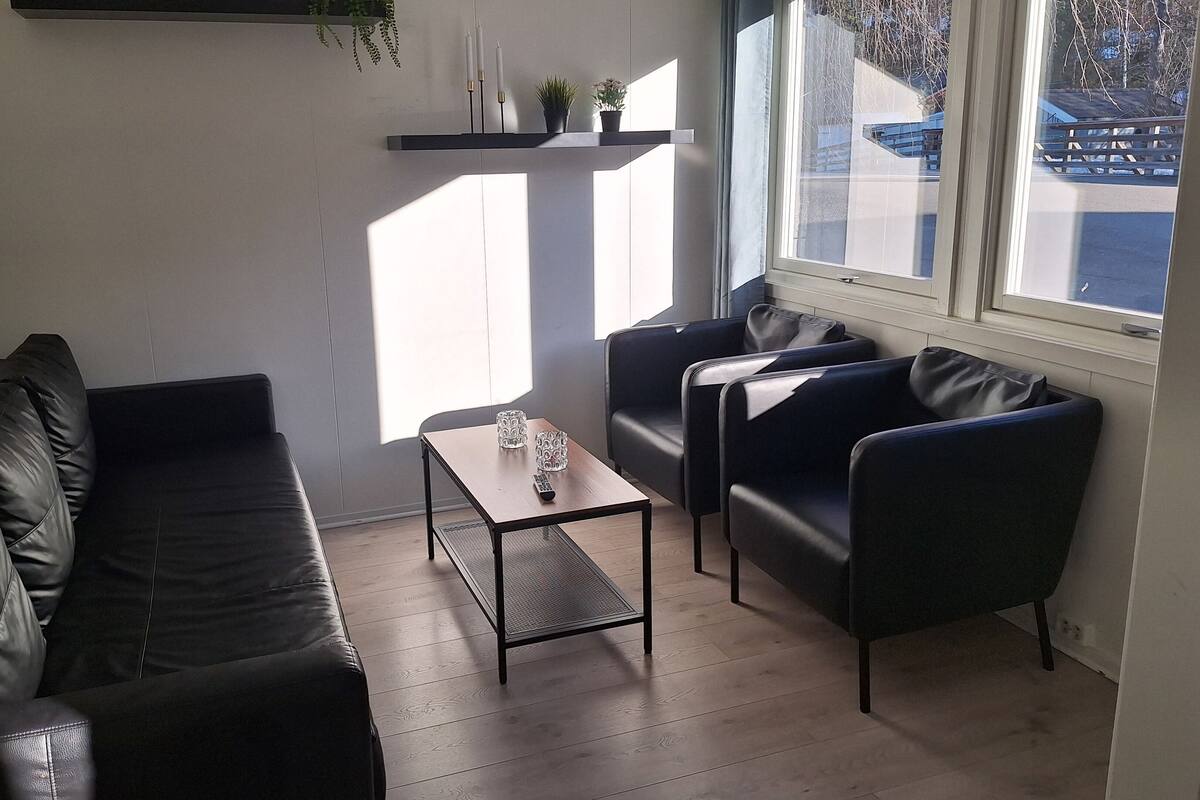 A modern living room with a leather sofa, a coffee table, and two armchairs. There is a window with a view of a building and a bridge.