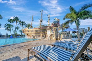 A pool area with lounge chairs, a ship-shaped structure, palm trees, and a clear sky. - 2BR Fireworks View With Pool Spa 5 Minutes to Disney (Orlando)