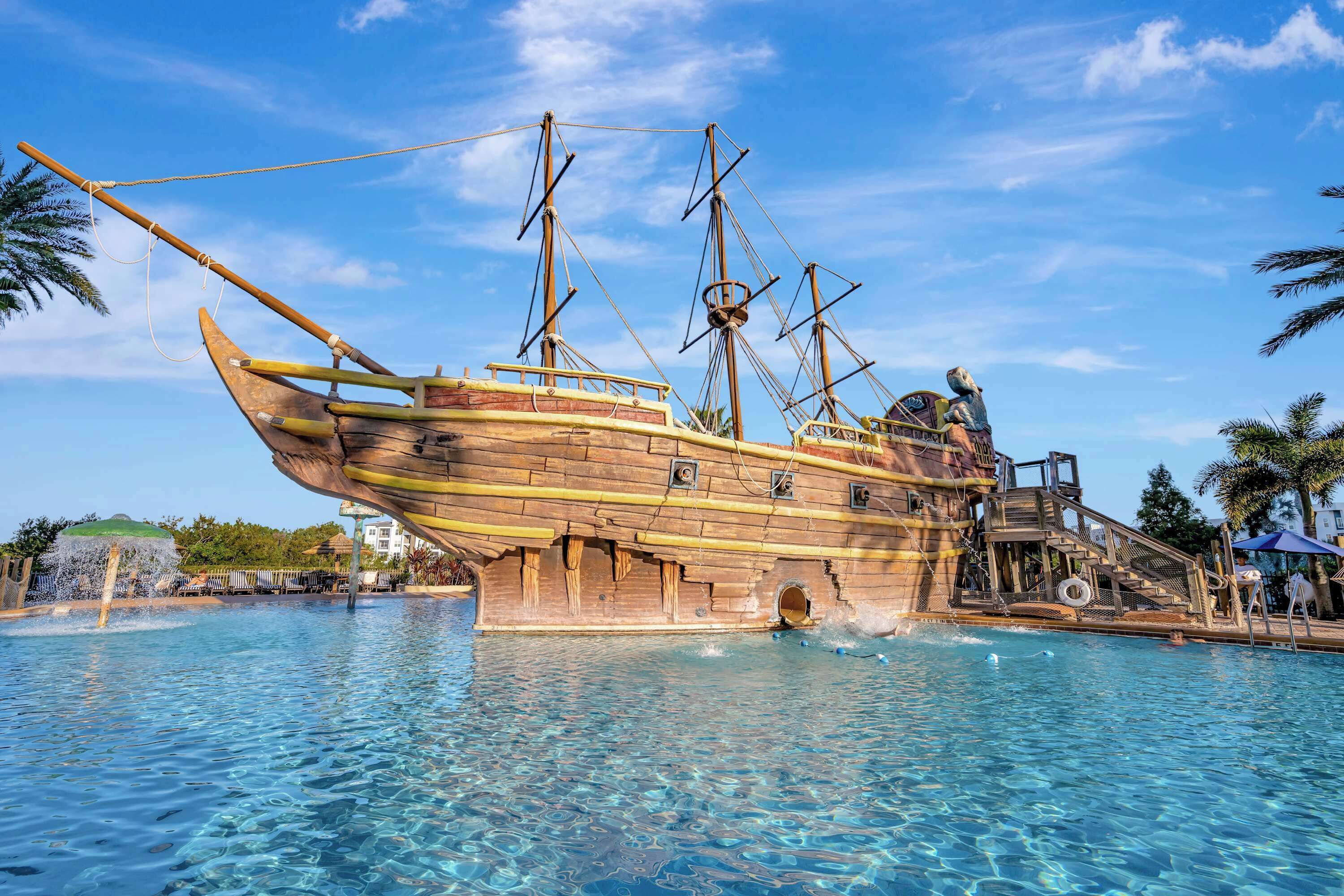 A large wooden pirate ship in a swimming pool, with a lifeboat and stairs leading up to it.