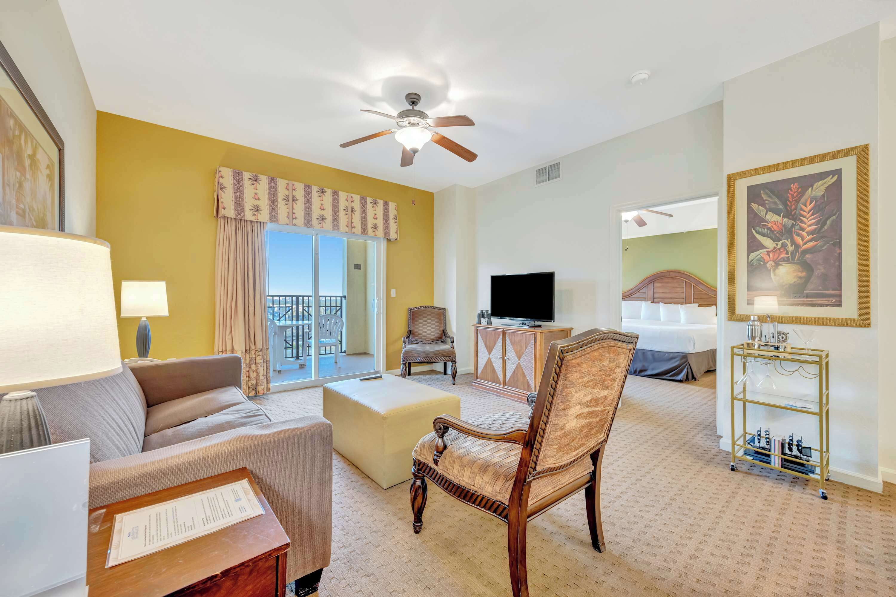 Habitación de hotel con sofá, sillón, reposapiés, televisor, ventilador de techo, armario y una pequeña mesa con un menú.