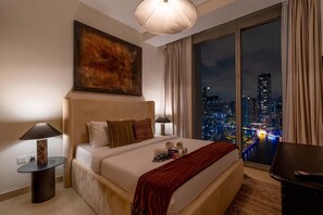 Ein modernes Hotelzimmer mit einem großen Bett, Blick auf die Stadt, einem Bild an der Wand und einem runden Tisch mit einer Lampe.