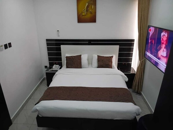A hotel room with a bed, a TV, a nightstand, and a painting on the wall. - Grand Churchill Luxury Hotel (Lagos)