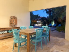 Ein Essbereich mit Holztisch und türkisfarbenen Stühlen, einer Holzskulptur an der Wand und Blick auf Pool und Palmen im Freien.