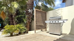 A barbecue grill in a patio area with a wooden deck, surrounded by palm trees and a woven screen. - Bright 3-Bedroom Villa Near Eagle & Palm Beach (Noord)