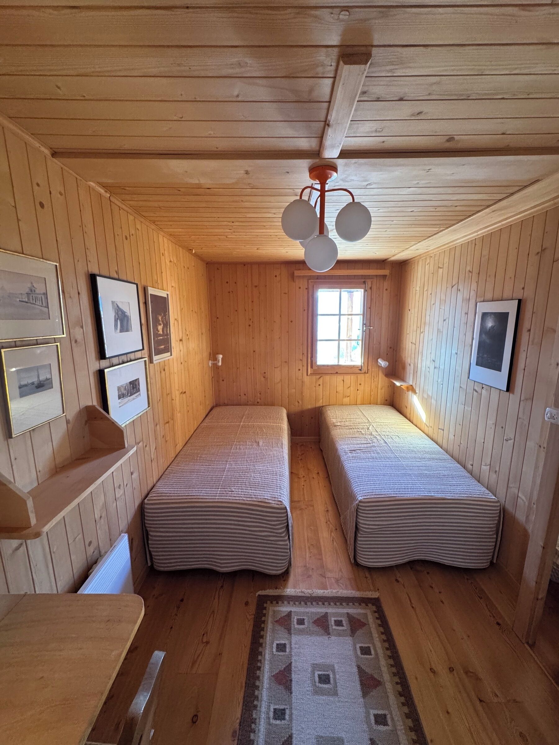 Une cabine avec deux lits simples, des murs en bois, un ventilateur de plafond, des tableaux encadrés et une fenêtre.