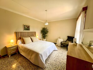 Une chambre à coucher comprenant un lit, une table de chevet, une lampe, une plante en pot, une chaise et une petite table ronde.