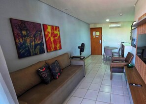 Ein Wohnzimmer mit einer Couch, zwei Bildern, einem Ventilator, einem Tisch mit Stühlen und einer Tür zum Essbereich.