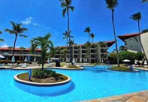Ein Resort mit einem zentralen Schwimmbecken, Palmen und einem mehrstöckigen Gebäude mit Balkonen.