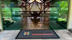 Entrada de hotel con puerta de cristal, felpudo con el nombre del hotel y cerca de madera con plantas.