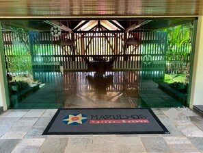 A hotel entrance with a glass door, a doormat with the hotel name, and a wooden fence with greenery. - Marulhos - Excelente Flat em Muro Alto (Ipojuca)