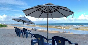 Ein Strand mit Reihen blauer Plastikstühle und weißen Sonnenschirmen, ein sandiger Uferstreifen und das Meer im Hintergrund.