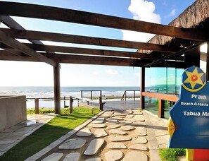 A beach access sign with a view of the ocean and a stone pathway leading to a covered area. - Marulhos - Excelente Flat em Muro Alto (Ipojuca)