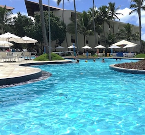 A swimming pool with lounge chairs and umbrellas, surrounded by palm trees and a building. - Marulhos - Excelente Flat em Muro Alto (Ipojuca)