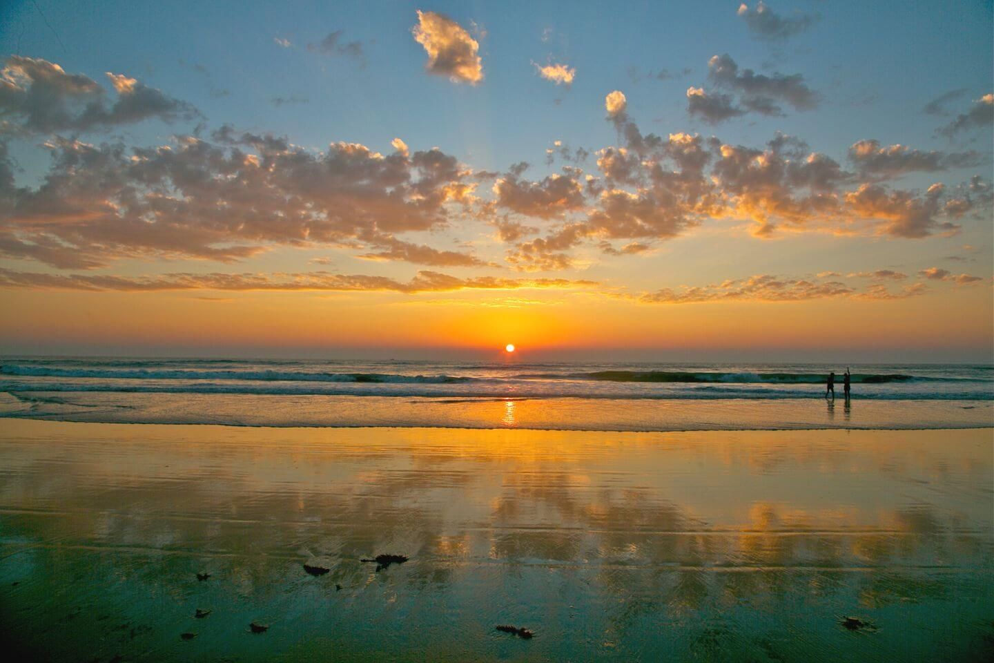 A beach scene with the sun setting over the ocean, two people standing on the shore, and birds flying over the water.