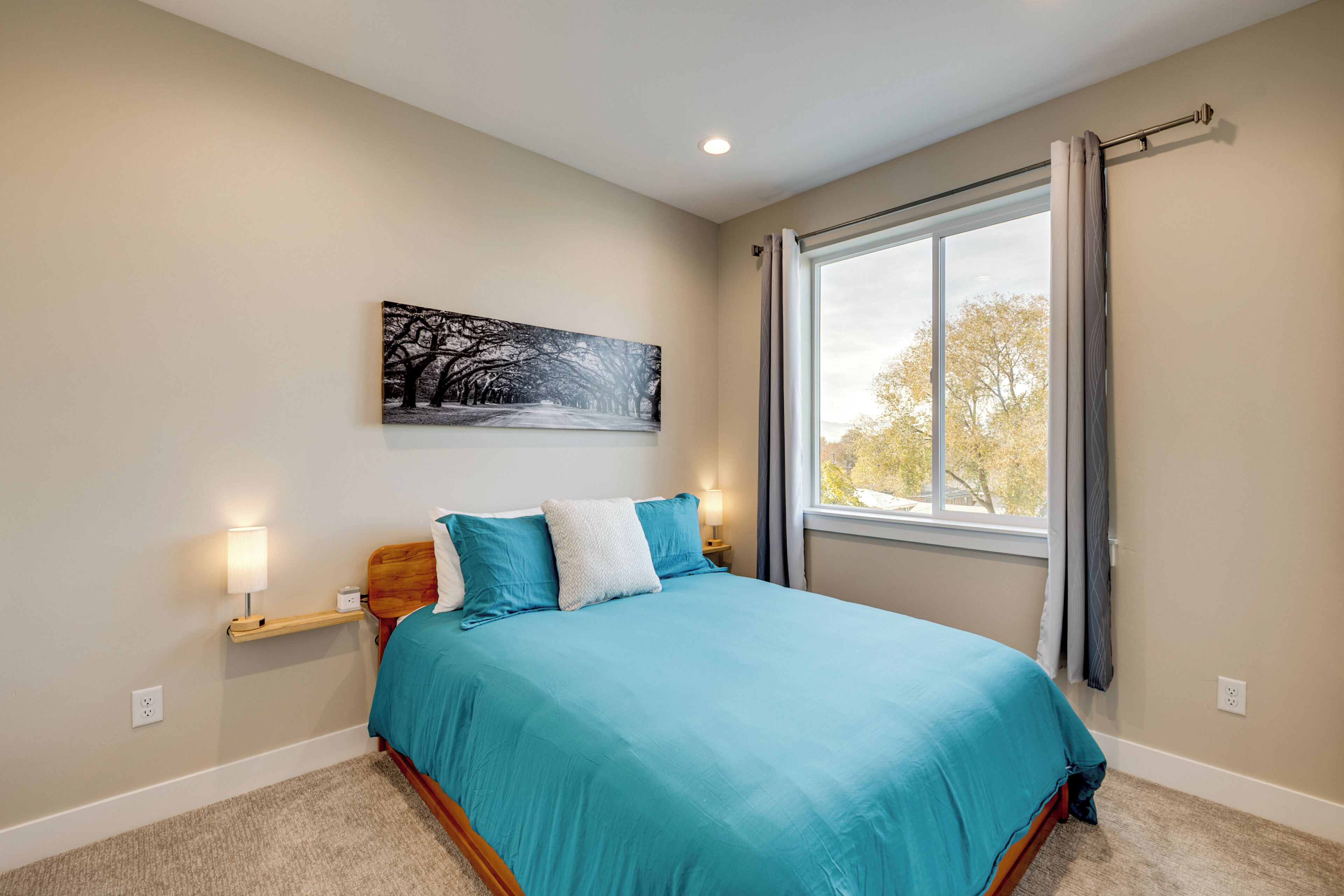 A bedroom with a large bed, a nightstand with a lamp, a wall-mounted painting, and a window with curtains.