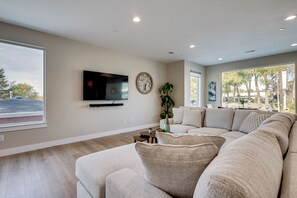 A modern living room with a large sectional sofa, a flat-screen TV mounted on the wall, and a large window offering a view of trees and a building. - Ski Resorts & SLC Fun! Brand New Millcreek Home (Salt Lake City)