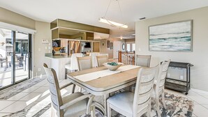 A modern dining area with a large table, chairs, and a kitchen in the background. - Canal-front Luxury Pool, Boat & Relax in Marco (Marco Island)