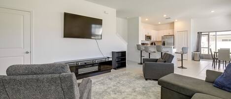 Ein modernes Wohnzimmer mit Fernseher, grauen Sofas, einem flauschigen Teppich und einer Küche im Hintergrund.