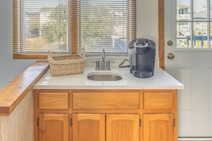 Una cocina con encimera blanca, armarios de madera, un lavamanos de acero inoxidable, una cafetera y una cesta de mimbre.