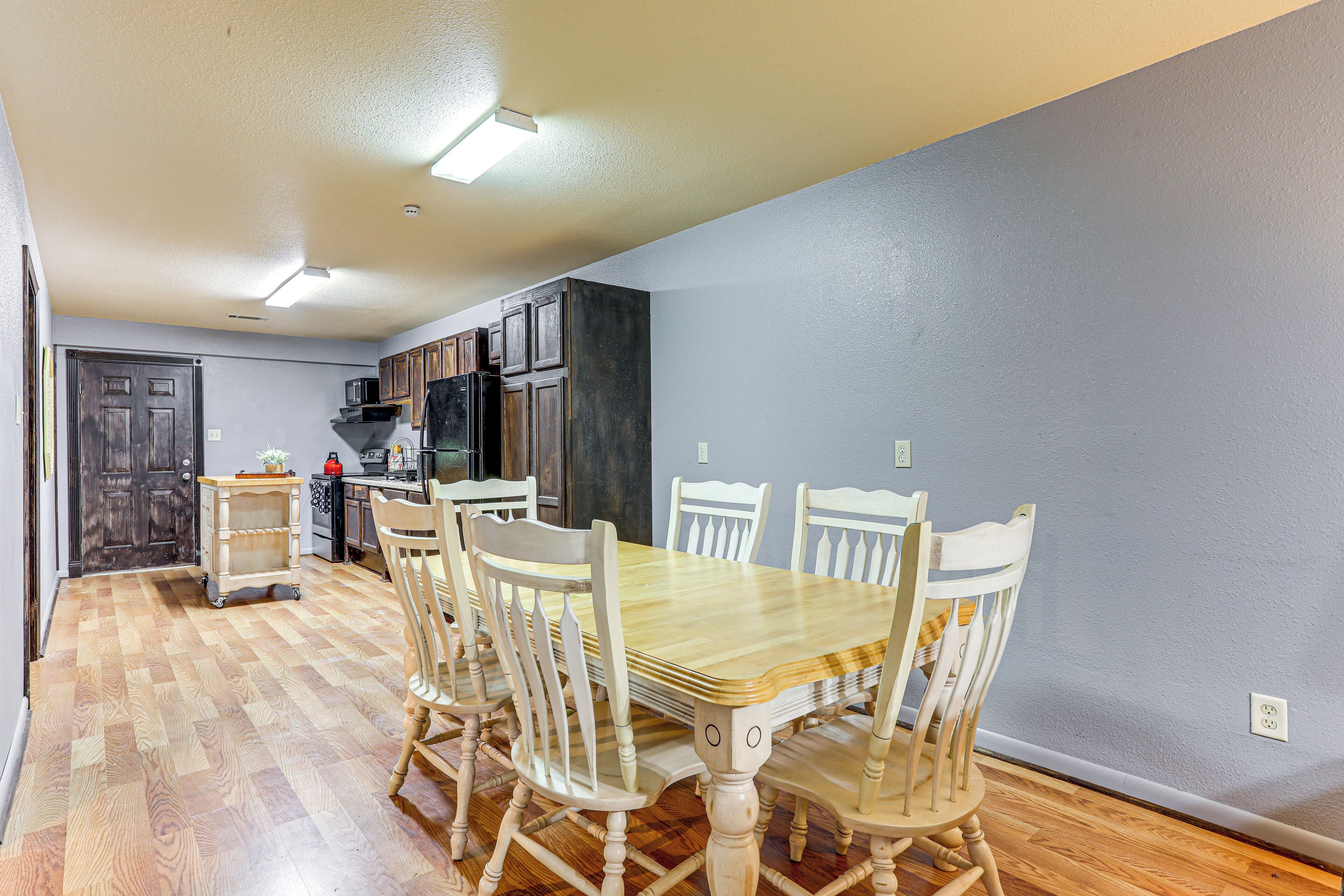 A dining area with a wooden table and chairs, a kitchen in the background, and a door leading to another room.