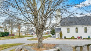 A residential street with a large leafless tree, a white house with green windows, and a stone bench in the foreground. - Nature Lovers Retreat By the High Trestle Trail! (Woodward)
