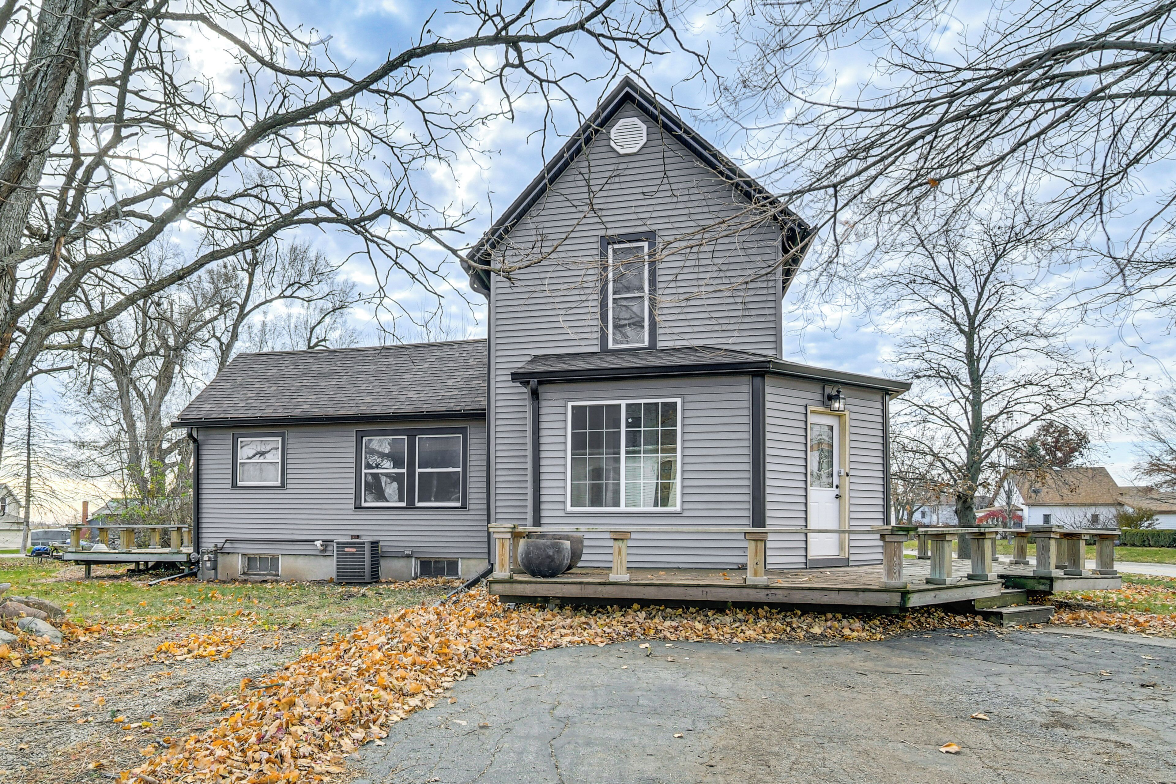 A two-story house with a porch and a small deck, surrounded by trees and a leaf-covered ground.