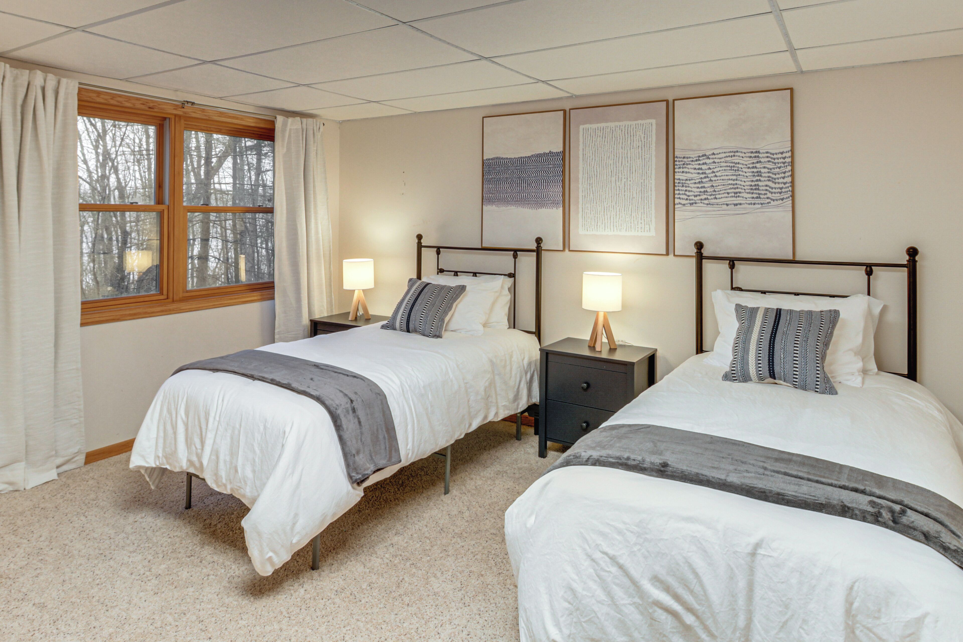 A bedroom with two beds, each with a bedside table and lamp, a window with curtains, and artwork on the wall.