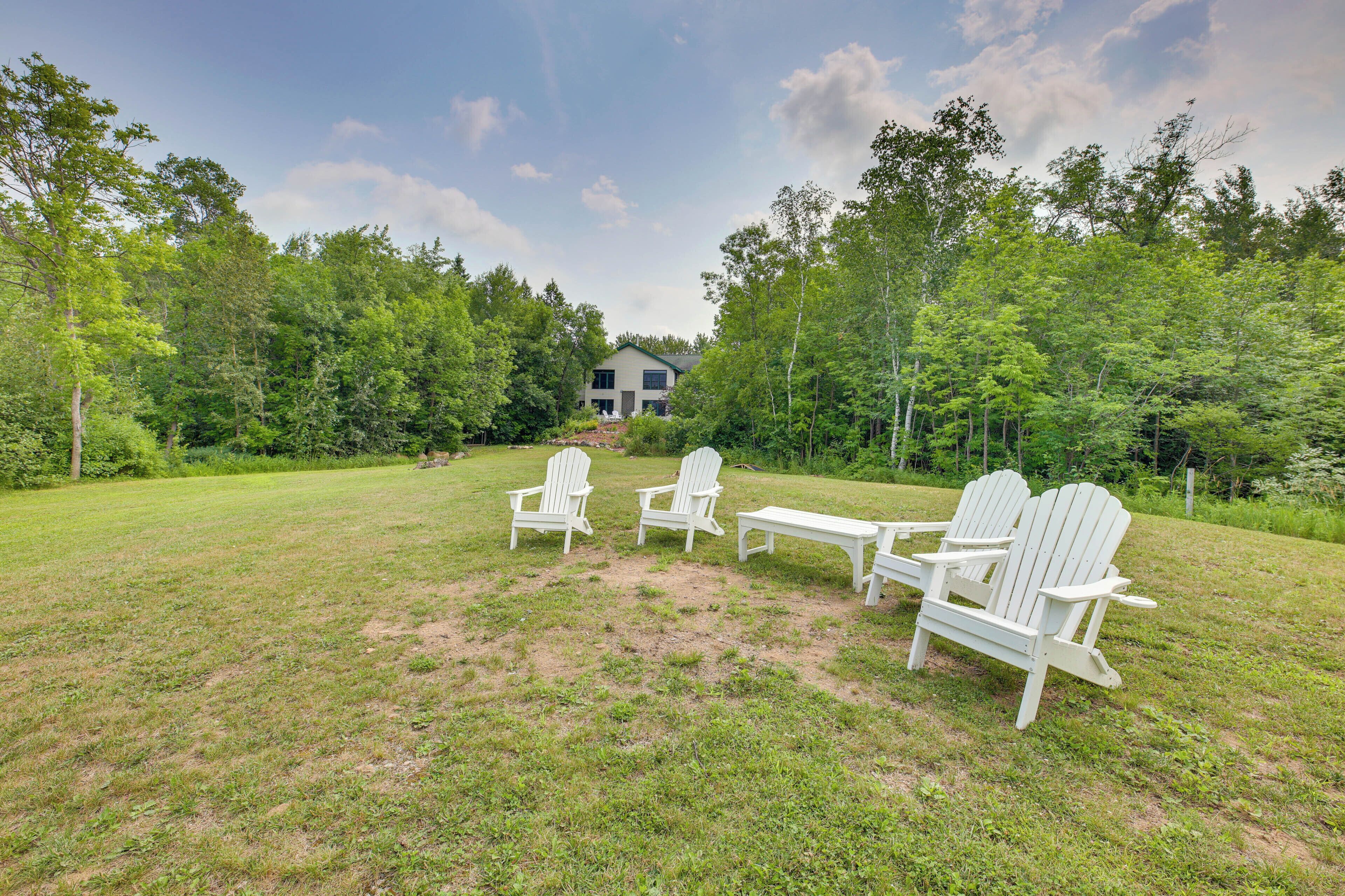 A grassy outdoor area with Adirondack chairs and a table, surrounded by trees and a house in the background.
