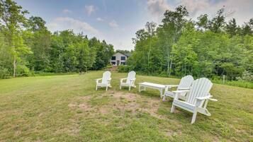 A grassy outdoor area with Adirondack chairs and a table, surrounded by trees and a house in the background.