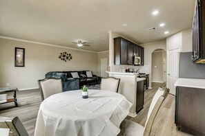 Ein modernes Wohnzimmer mit einem runden Esstisch, dunklen Holzschränken und einer Küchenzeile, ebenfalls mit dunklen Holzschränken und weißen Arbeitsplatten.