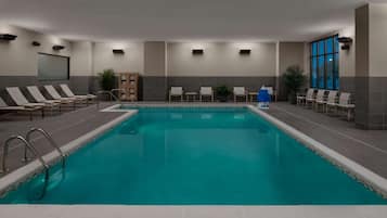 Una piscina con sillas de descanso y una mesita, rodeada de muros con ventanas y suelo de baldosas.