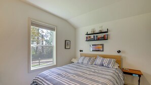 Un dormitorio con un ventanal grande, una cama con ropa de cama a rayas, una mesita de noche y un estante con libros y objetos decorativos.