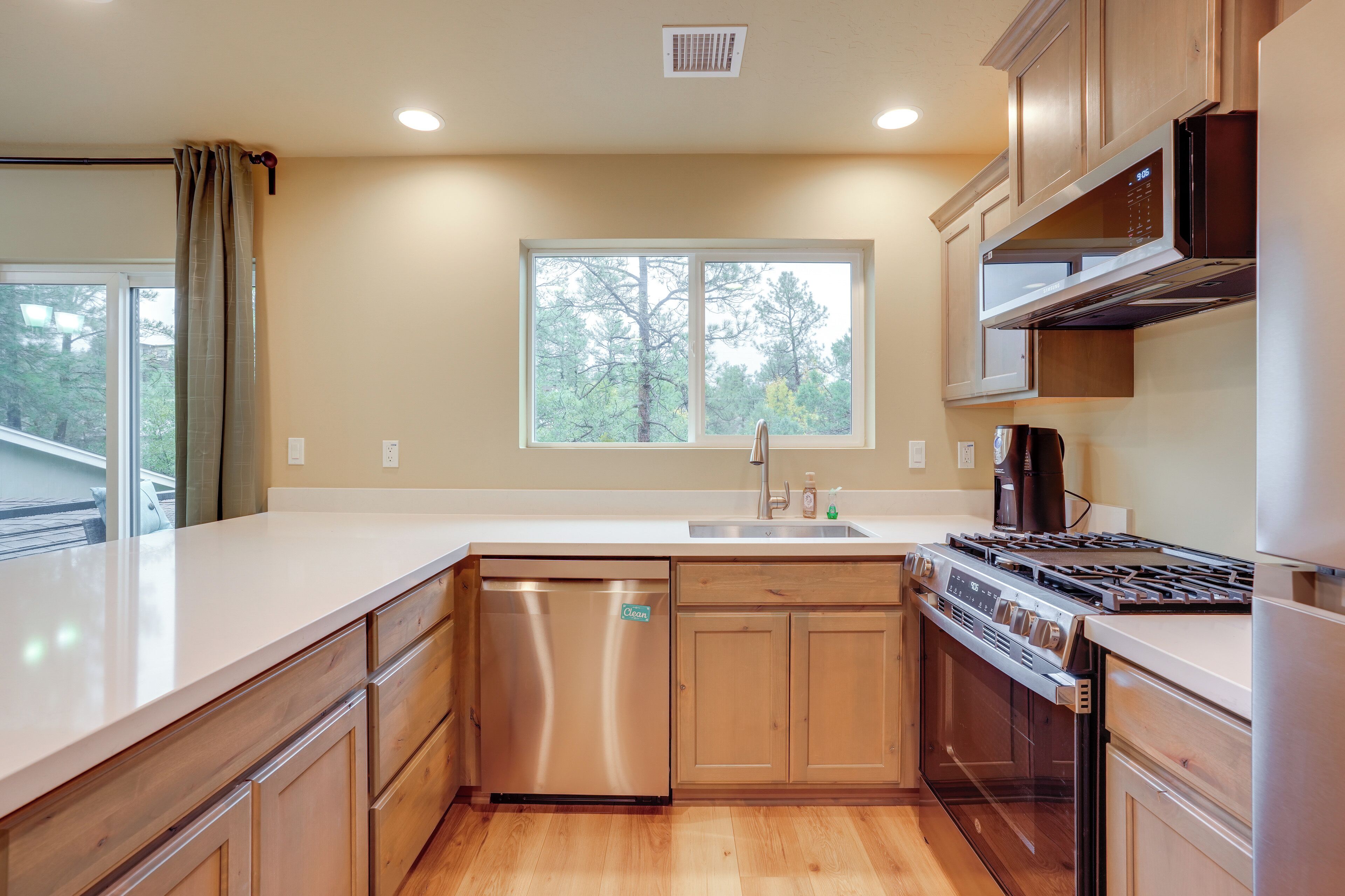A modern kitchen with wooden cabinets, a stainless steel dishwasher, a built-in oven, and a window providing a view of trees.