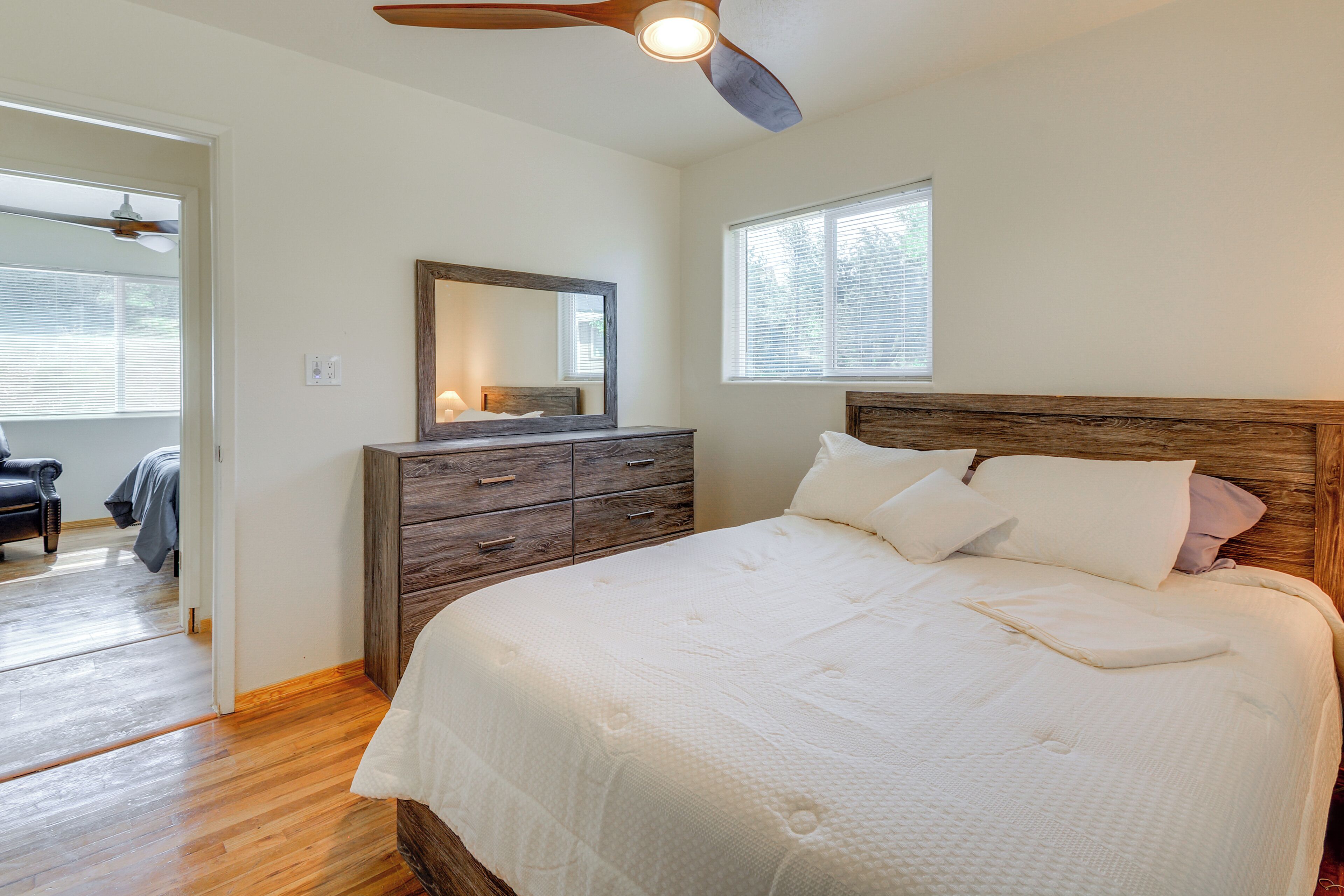 Een slaapkamer met een houten hoofdbord, een groot bed met wit beddengoed, een dressoir met lades, een spiegel en een plafondventilator.