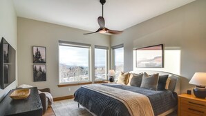A bedroom with a bed, a desk, a chair, a ceiling fan, and large windows offering a city view. - Luxe Home w/ Hot Tub and Views in Winter Park! (Winter Park)