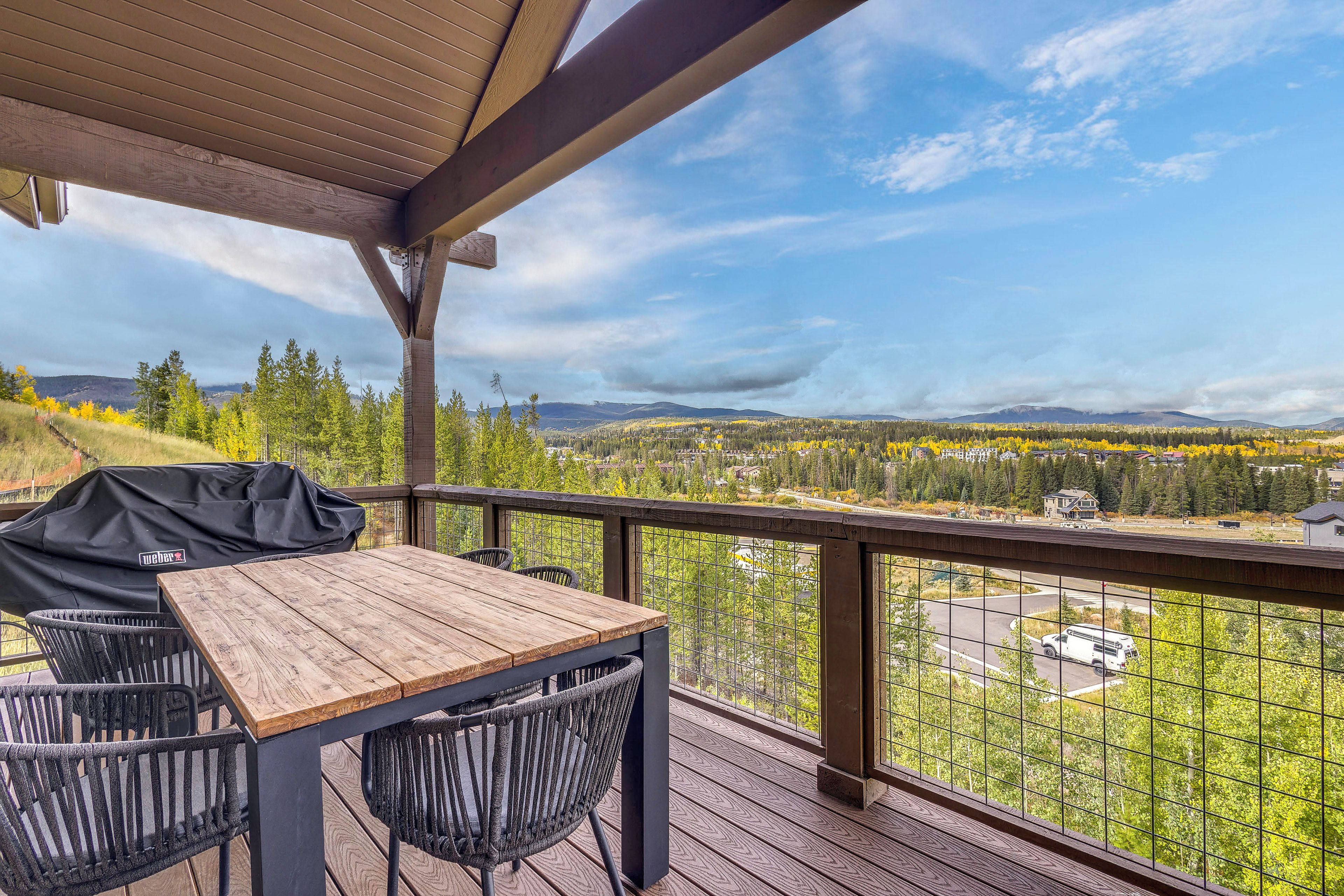 A wooden deck with a table and chairs, overlooking a scenic landscape with trees and mountains.