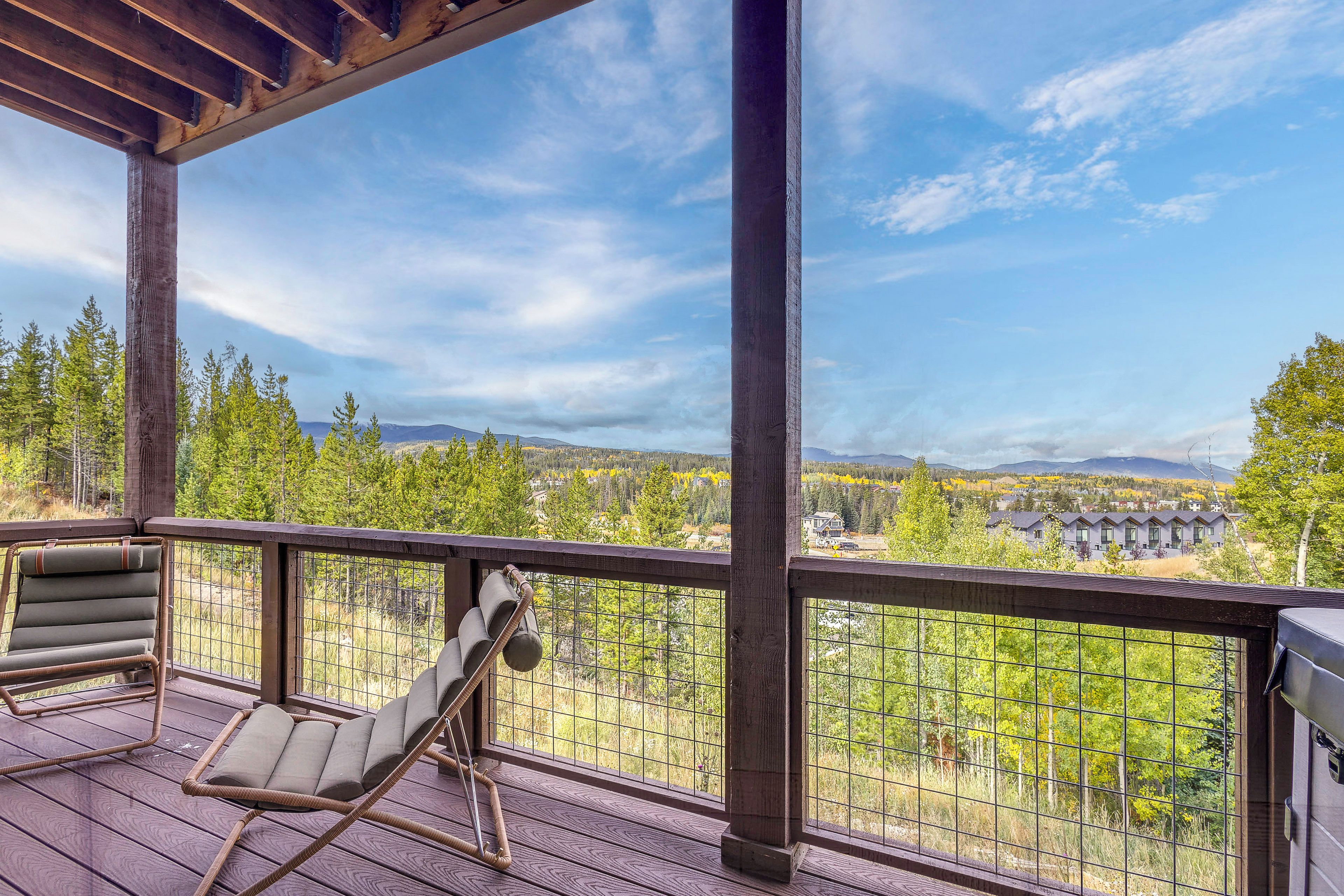 Une terrasse en bois offrant une vue sur une forêt et des montagnes. On y trouve des chaises en osier et une rambarde en métal.