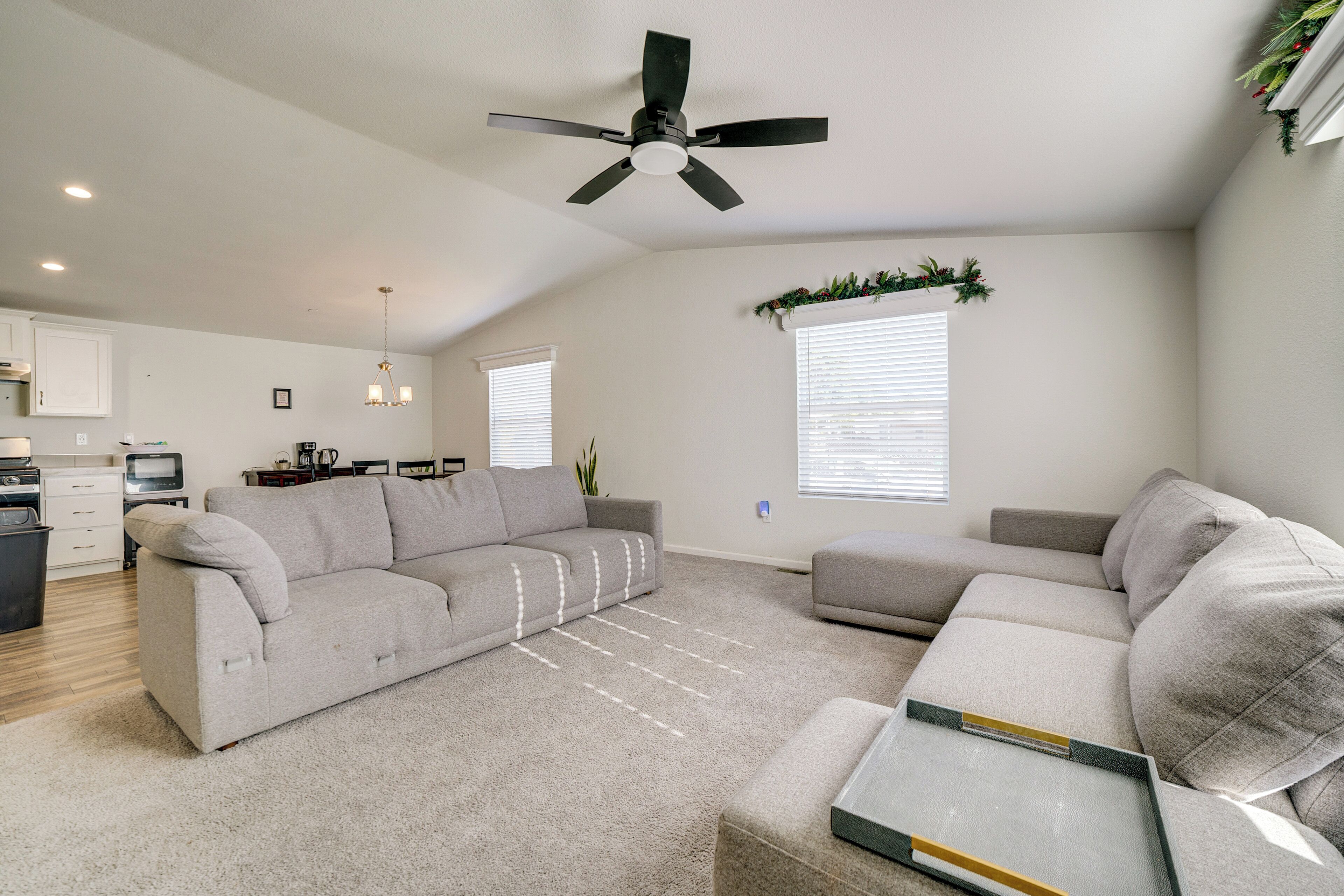 A living room with a grey sectional sofa, a small ottoman, a ceiling fan, and a kitchen area in the background.