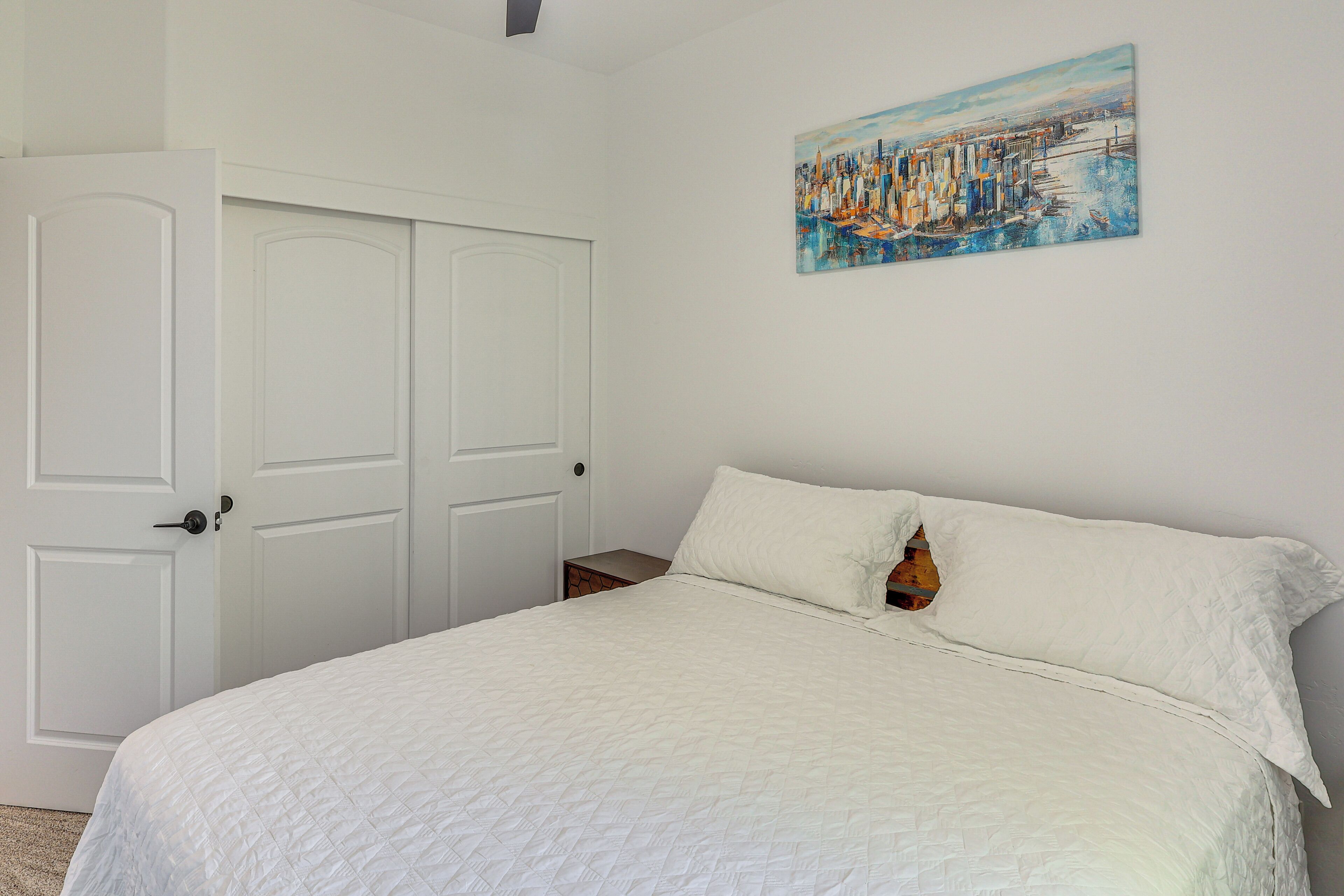 A bedroom with a white bed, a door, and a painting on the wall.