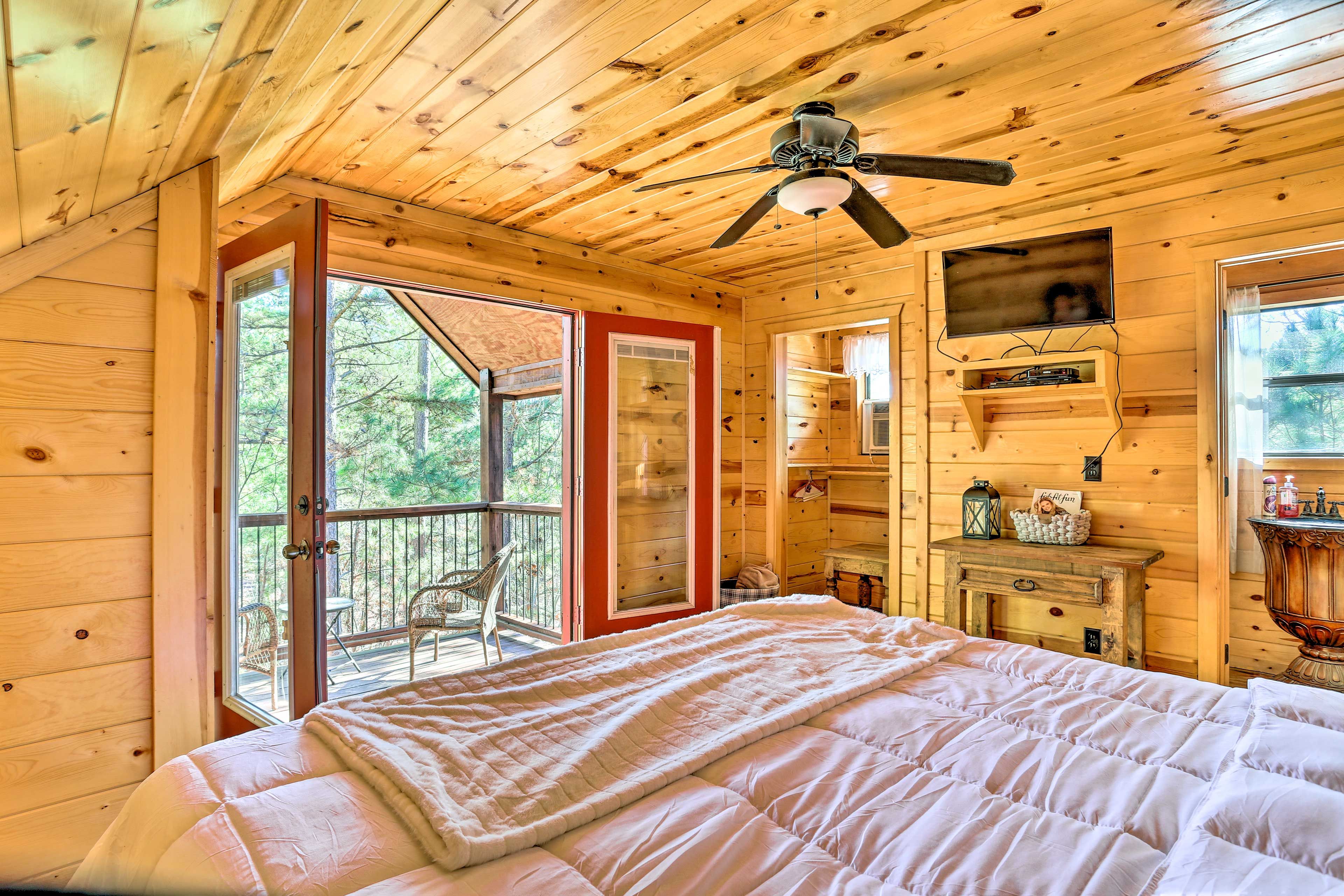 A wooden cabin bedroom with a bed, a ceiling fan, a small table, and a window overlooking a deck.