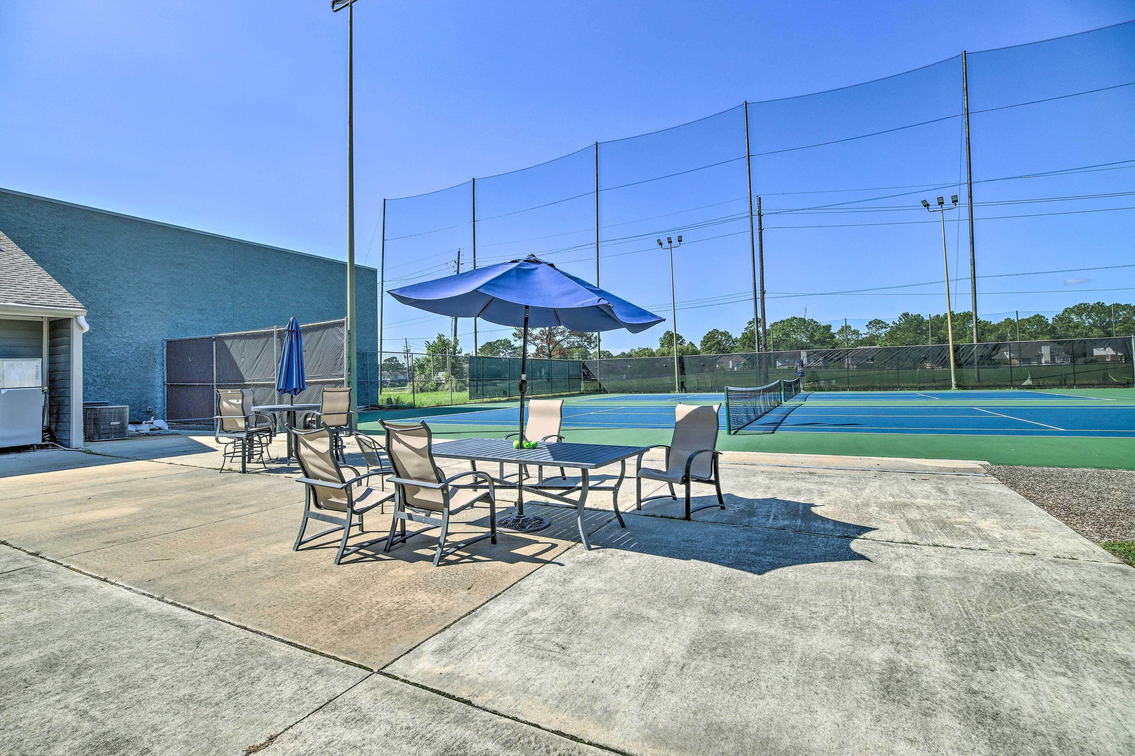 An outdoor patio with a table and chairs, a blue umbrella, and a tennis court in the background.