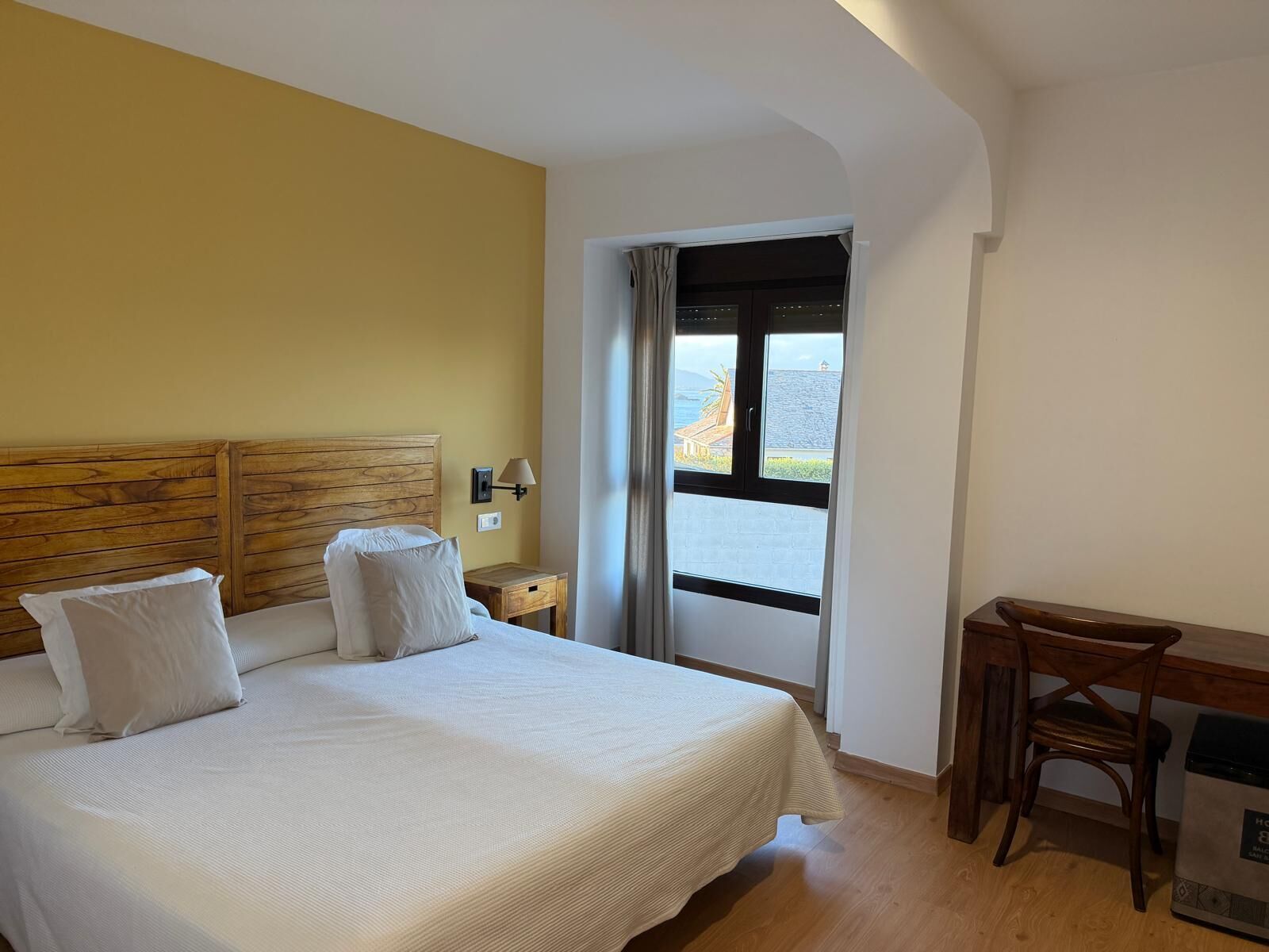 A hotel room with a bed, a desk, and a window with a view of the sea.