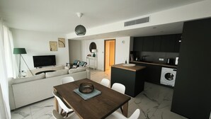 Ein zeitgemäßer Wohnraum mit Essbereich, einer Couch, einem Fernseher, einer Lampe und einer Küche inklusive Waschmaschine.